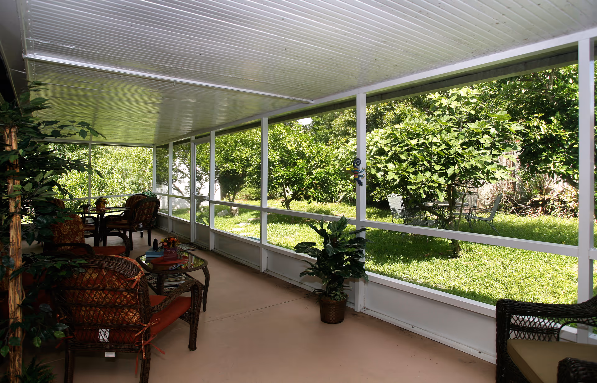 A covered screened porch with wicker chairs and tables, overlooking a green garden with trees and outdoor metal chairs and table set.