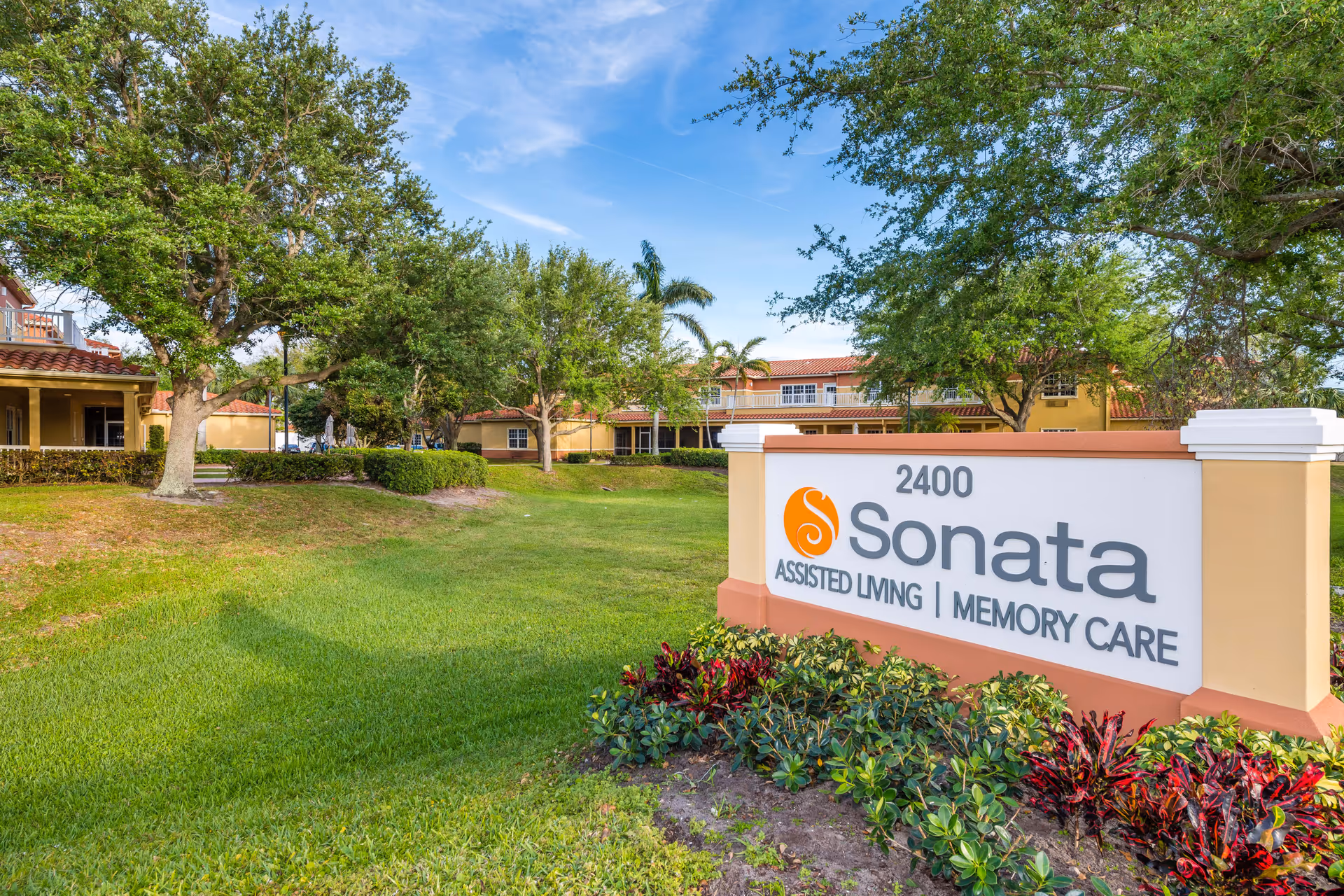 Outdoor view of Sonata by AgeWell Solvere Living facility with a large sign displaying the name and services offered, surrounded by green grass, trees, and landscaping under a blue sky.