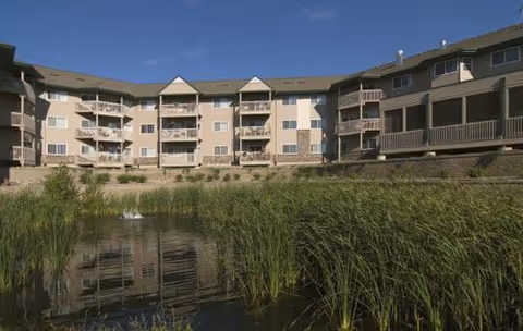 Exterior view of a multi-story senior living building with balconies reflected in a pond edged by reeds.