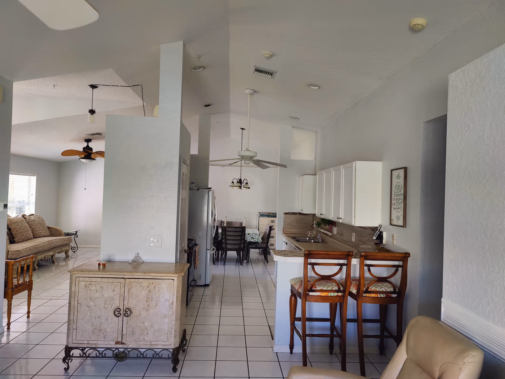 Interior view of a senior living facility showing a kitchen area with white cabinets, a countertop with two wooden bar stools, and a dining area with a table and chairs in the background. To the left, there is a living room area with a cushioned bench and a ceiling fan. The floor is tiled with white tiles, and the walls are painted light gray.