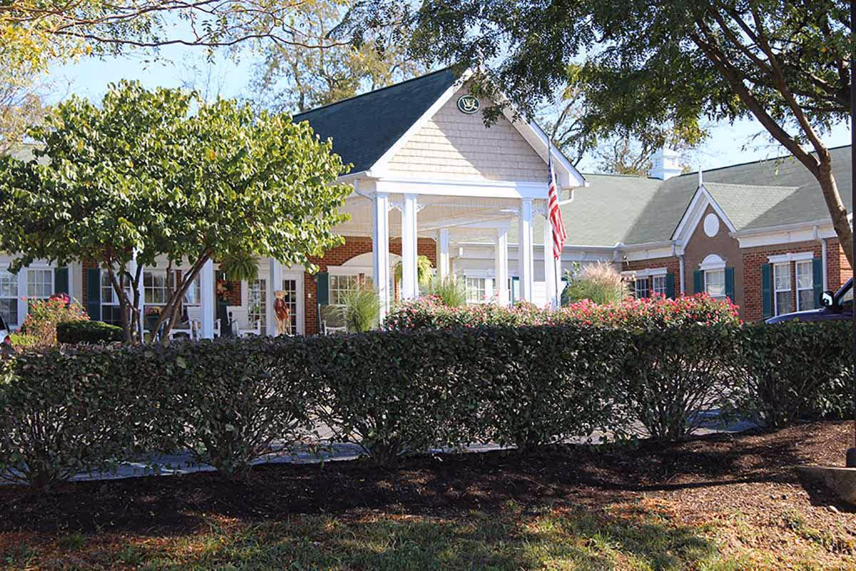 Exterior view of Windsor Gardens Senior Living facility showing a brick building with white columns at the entrance, green shutters on windows, an American flag, and well-maintained landscaping with bushes, trees, and flowers.