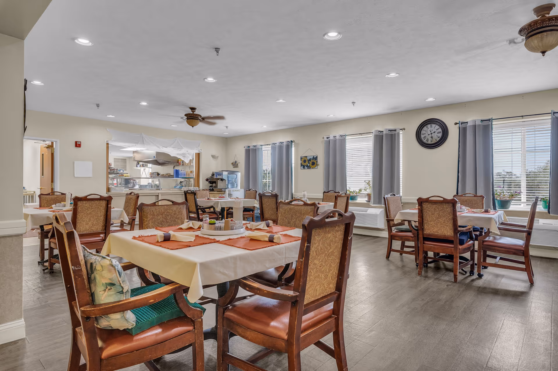 A spacious dining room in a senior living facility with several tables covered in white and orange tablecloths, each set with napkins and condiments. The room has wooden chairs with cushioned seats and backs, large windows with gray curtains, a wall clock, and a ceiling fan. In the background, a kitchen area is visible through a serving window.