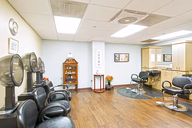 Interior view of a salon area with multiple black salon chairs, hair drying stations, and a hair washing sink. The room has wooden flooring, light-colored walls, ceiling lights, and a small wooden cabinet with decorative items.