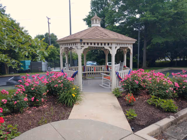 A white wooden gazebo with a brown shingled roof is centered at the end of a concrete pathway. The gazebo is surrounded by vibrant pink flowers and green shrubs on either side. There are white rocking chairs inside the gazebo, and trees and a parking lot are visible in the background.