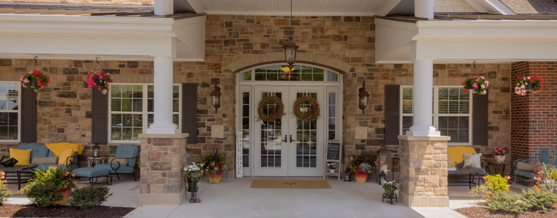 Entrance of Bickford of Suffolk facility featuring double glass doors with wreaths, stone walls, white pillars, hanging flower baskets, outdoor seating with cushions and pillows, and various potted plants and decorations.