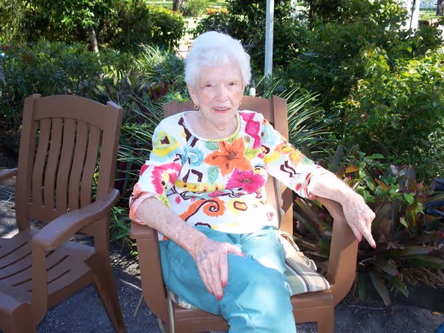 An elderly woman with white hair sitting outdoors on a brown plastic chair surrounded by green plants and bushes. She is wearing a colorful floral top and teal pants, smiling at the camera.
