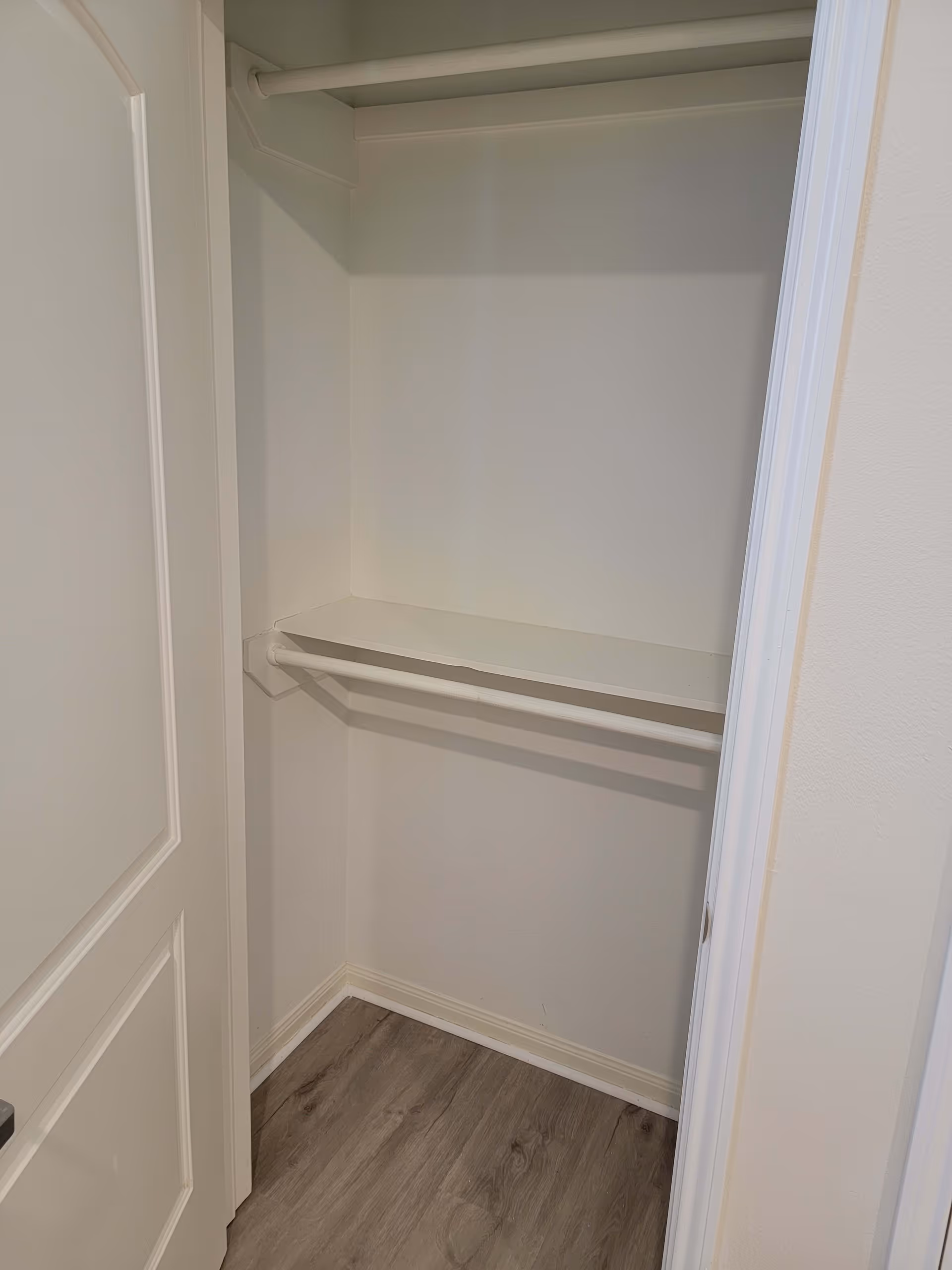 Empty white closet with a hanging rod and a shelf above it, wooden floor, and a partially open white door.