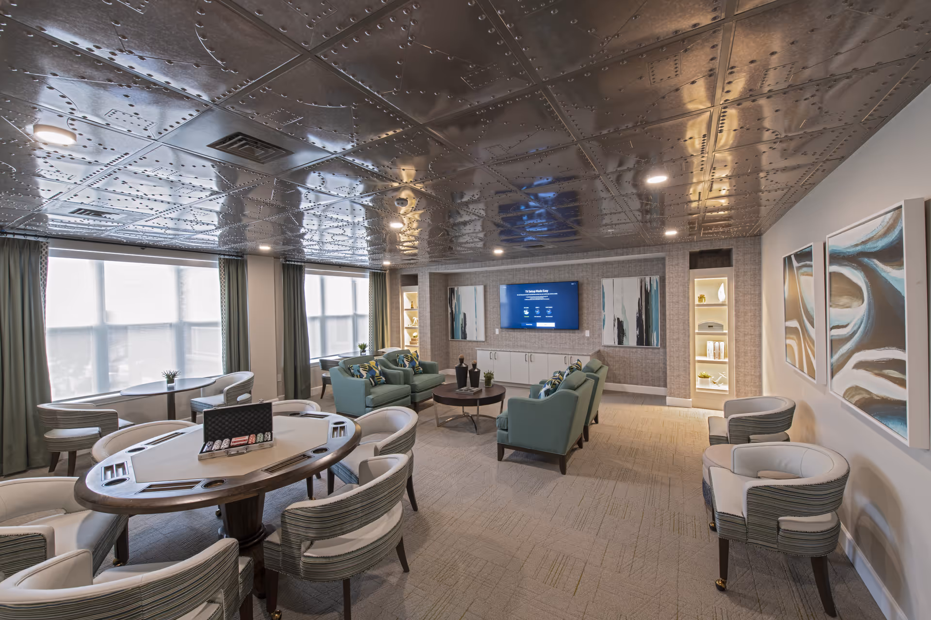 A modern senior living facility common room with a metallic riveted ceiling, a poker table with chairs, several armchairs arranged around a round coffee table, a wall-mounted TV, built-in shelves with decorative items, large windows with curtains, and abstract artwork on the walls.