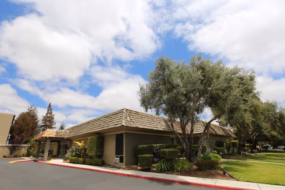 Front exterior of a single-story senior living building with a shingled roof, landscaped shrubs and trees under a partly cloudy sky.