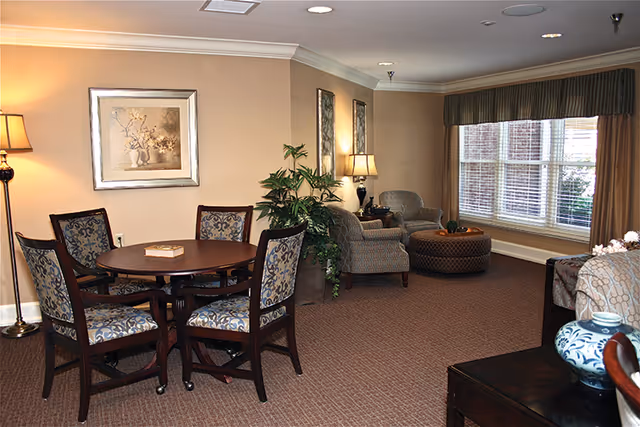 A cozy living room area with a round wooden table surrounded by four upholstered chairs with patterned fabric. There are two armchairs and a large ottoman near a window with blinds and curtains. The room is decorated with framed artwork, a floor lamp, a table lamp, and green plants. The carpet is brown, and the walls are painted beige.