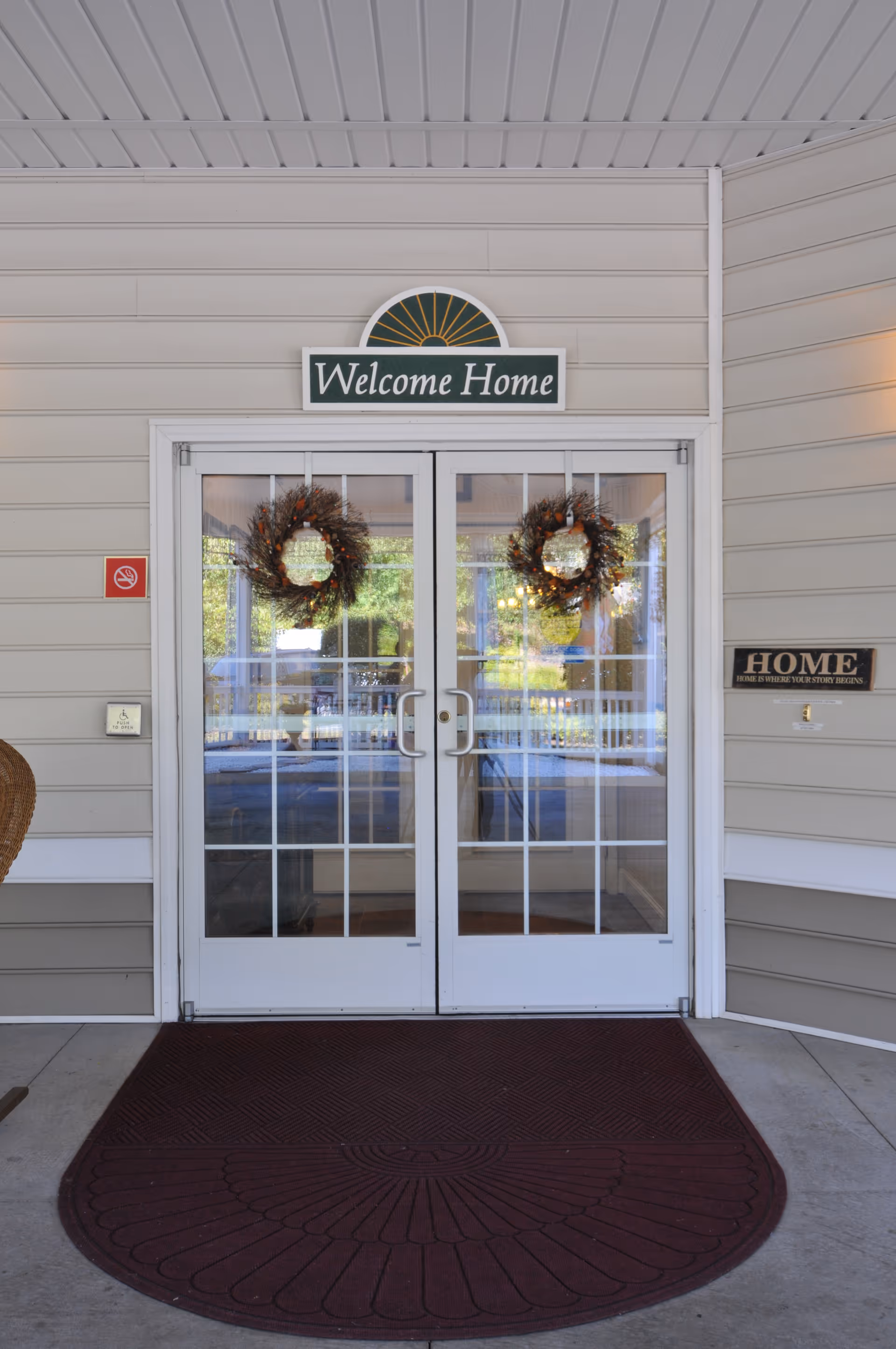Glass double entrance doors decorated with wreaths under a 'Welcome Home' sign at the front of a senior living facility.