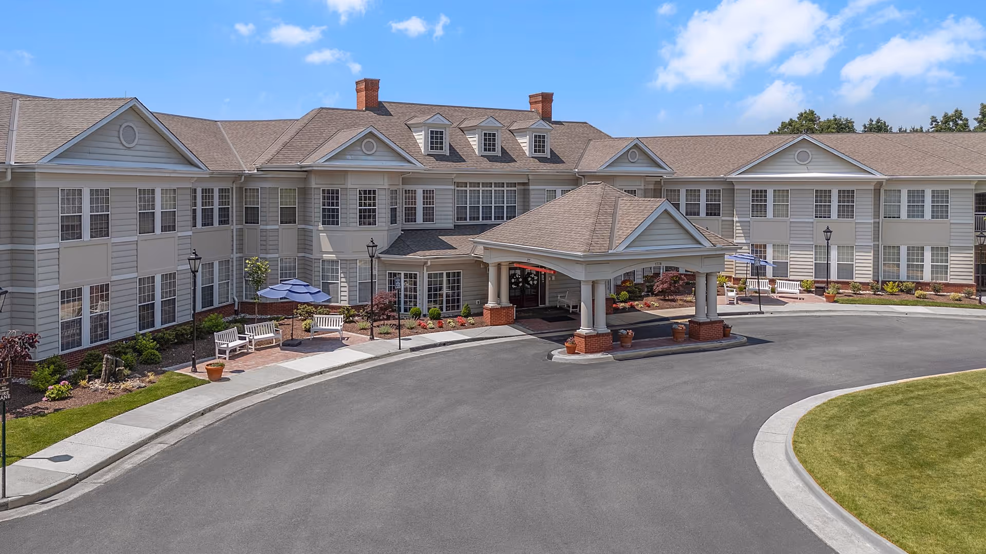 Front entrance of a two-story senior living building with a covered porte-cochère, circular driveway, benches and landscaping under a blue sky.