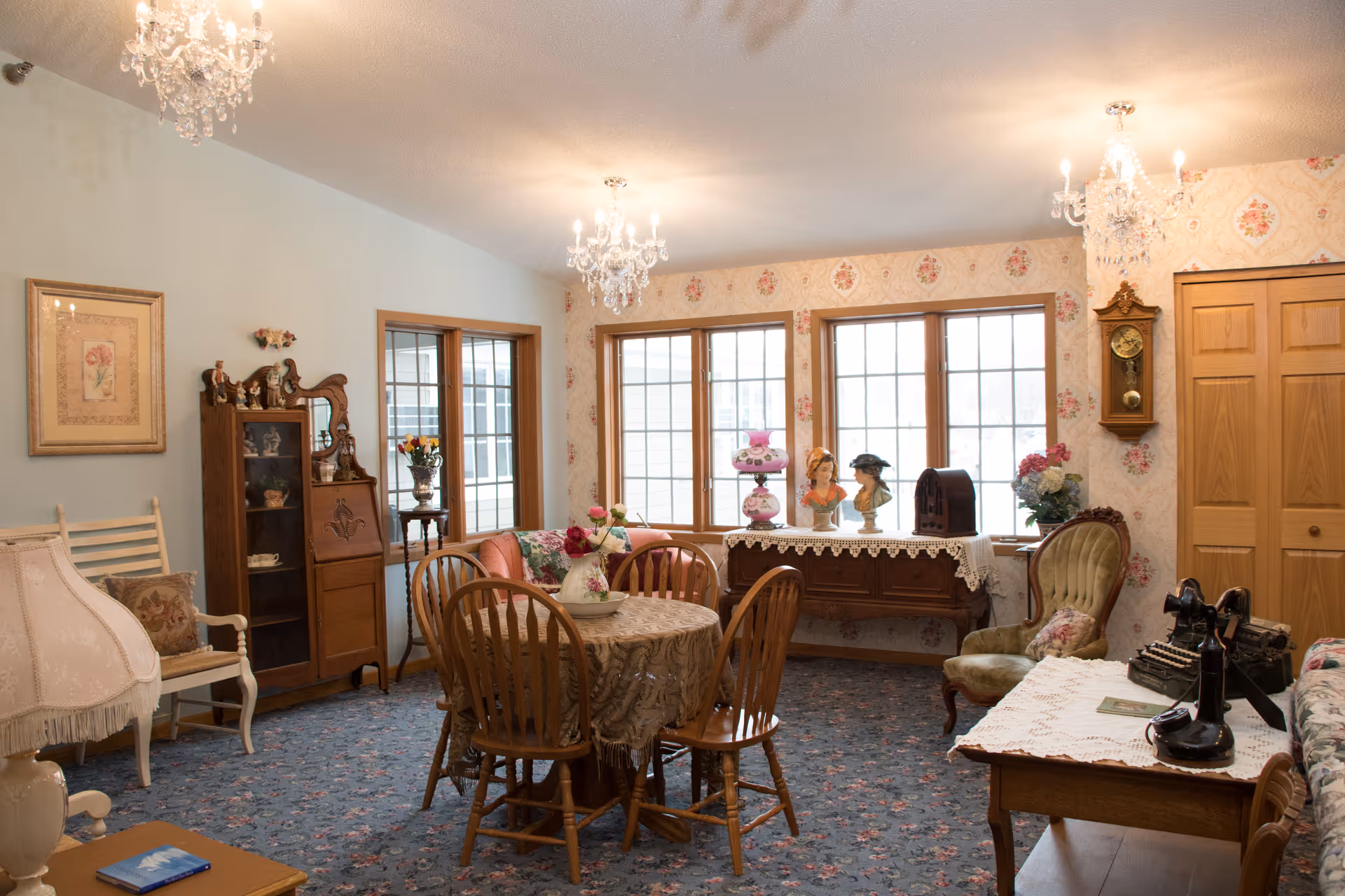 Bright vintage-style senior living common room with a round dining table and wooden chairs, upholstered seating, and decorative cabinets under large windows.