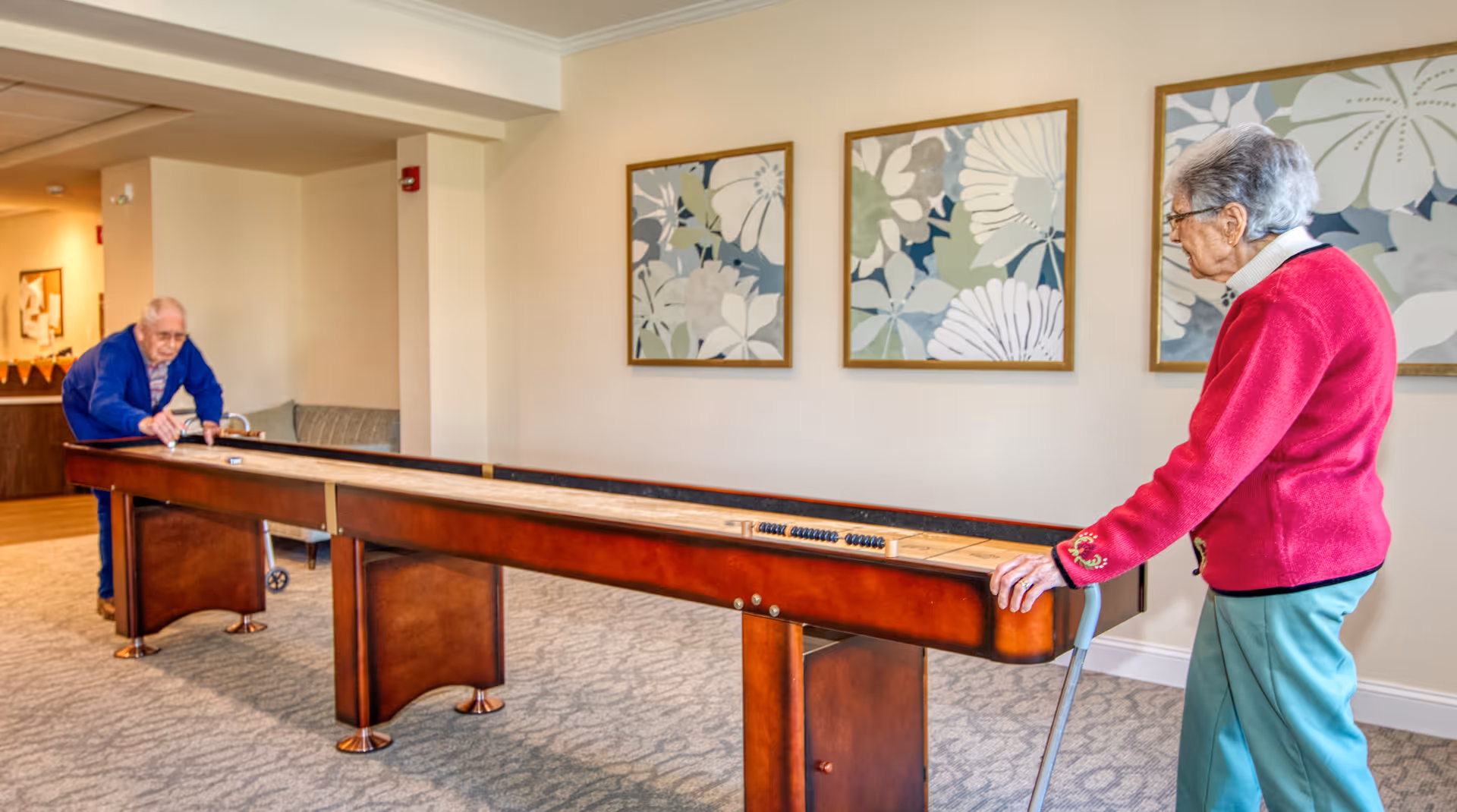 Two elderly individuals playing shuffleboard indoors in a well-lit room with carpeted floor and floral artwork on the wall.