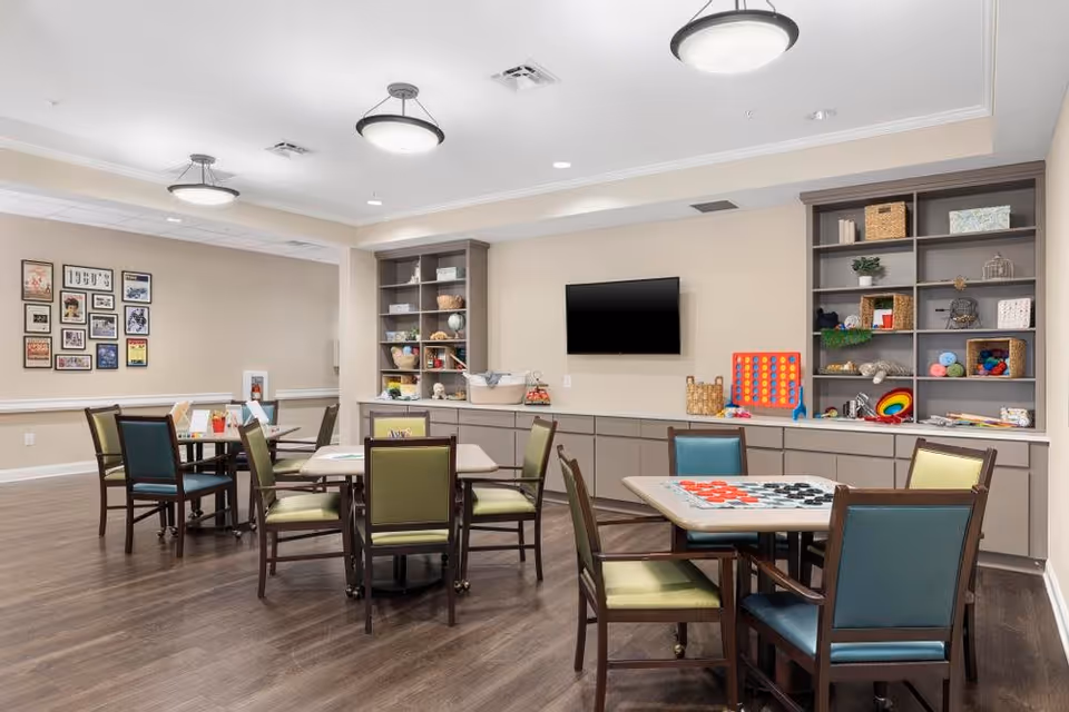 A well-lit activity room with multiple tables and chairs arranged for group activities. The tables have board games like checkers and Connect Four set up. There are built-in shelves along the back wall filled with various games, books, and decorative items. A flat-screen TV is mounted on the wall above the cabinets. The room has wooden flooring and neutral-colored walls with framed pictures on one side.
