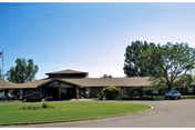 Exterior view of a single-story building with a covered entrance, surrounded by trees and a well-maintained lawn under a clear sky.