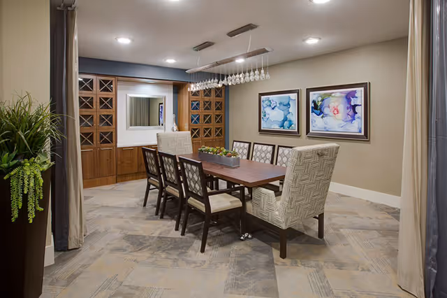 Communal dining room with a long wooden table surrounded by upholstered chairs, pendant lighting, wall art, and built-in cabinetry.