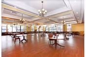 Large bright dining room with round wooden tables and chairs, chandeliers, and large windows along the far wall.