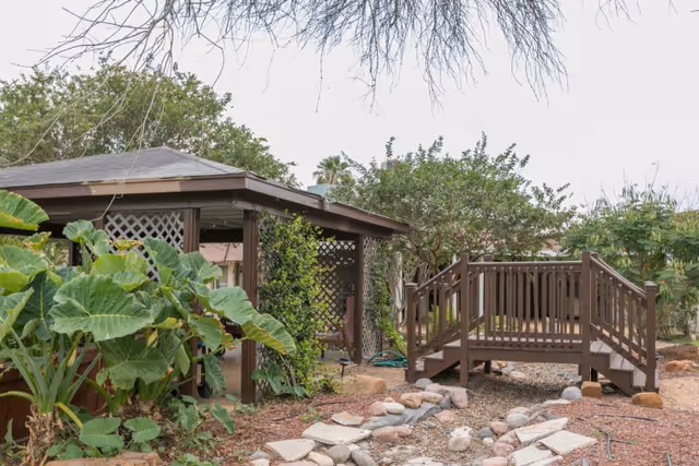 Outdoor garden area with a wooden gazebo covered in greenery, a small wooden bridge over a dry stone path, surrounded by various plants and trees under a cloudy sky.