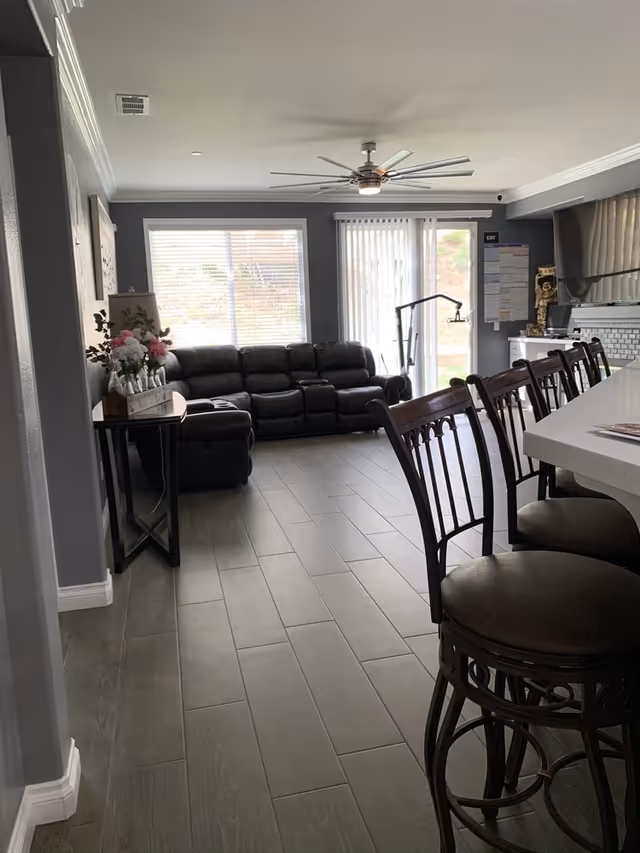 Interior view of a living room area with a dark leather sectional sofa against the wall, a ceiling fan with lights, large windows with blinds, a row of wooden bar stools with cushioned seats along a white countertop, and a small table with a flower arrangement on the left side.