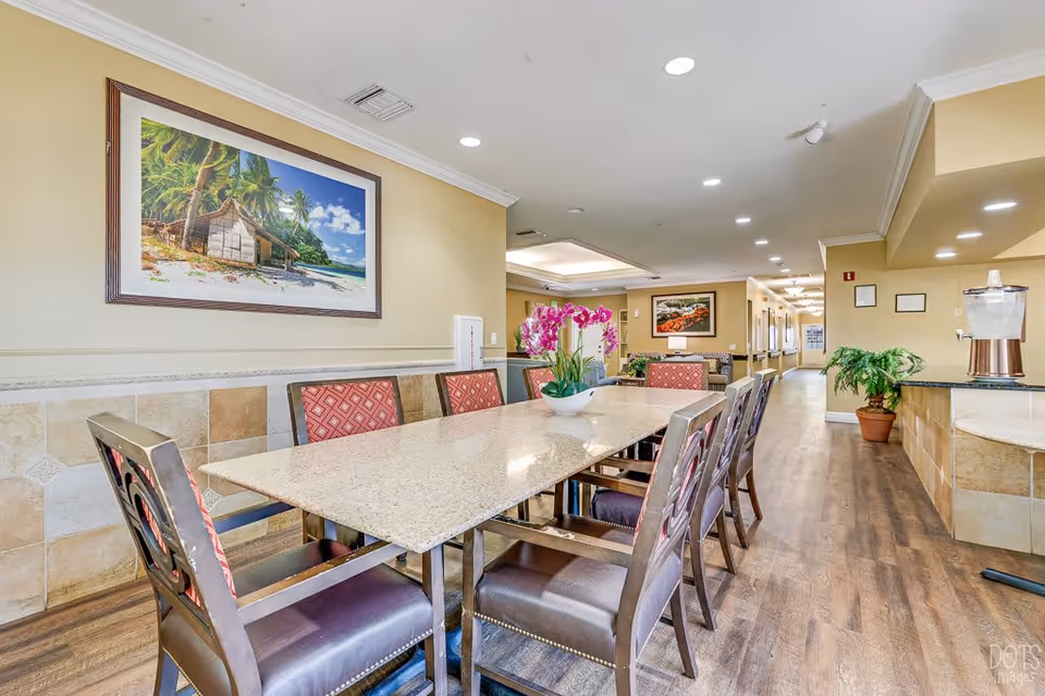 A dining area in a senior living facility with a long rectangular table surrounded by eight chairs. The table has a decorative flower arrangement in the center. The room has light-colored walls with a large framed beach-themed painting on one wall. The floor is wooden, and there is a water dispenser on a counter to the right. The space is well-lit with recessed ceiling lights and has a hallway leading further into the facility.