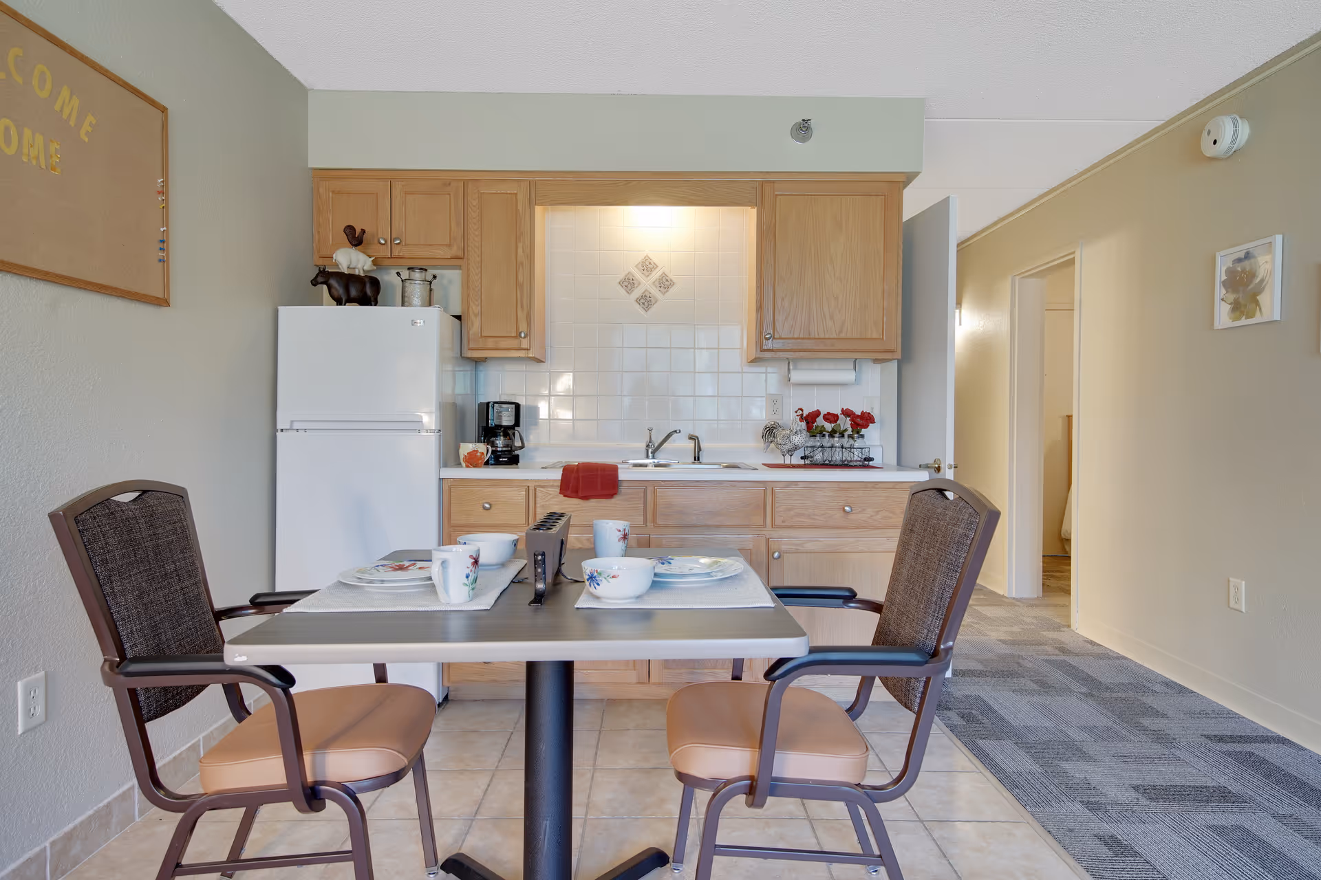 A small kitchen and dining area in a senior living facility. The kitchen has wooden cabinets, a white refrigerator, a coffee maker, and a sink with a red towel hanging on the counter. A dining table set for two with cups, bowls, and plates is in the foreground, with two cushioned chairs. The walls are light-colored, and there is a hallway leading to another room on the right side.