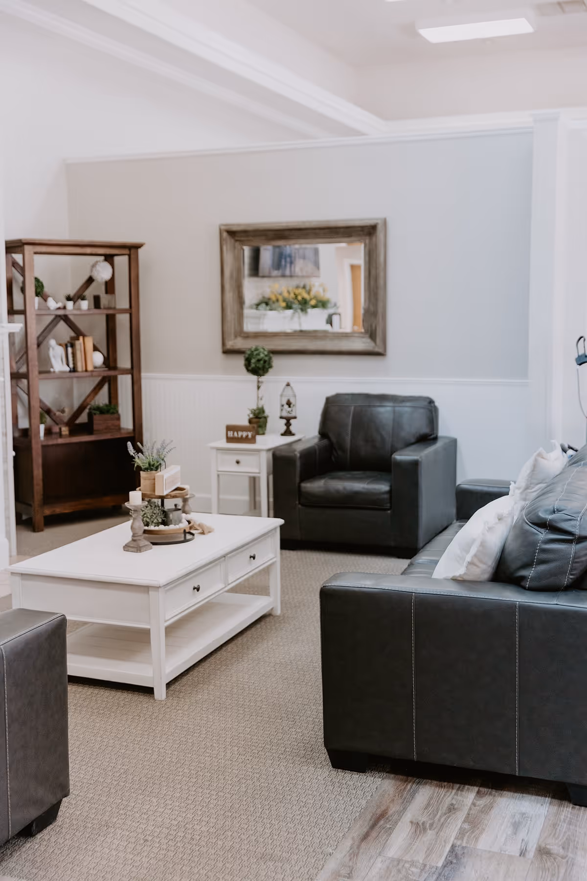A cozy living room area with dark leather armchairs and a sofa, a white coffee table with decorative items, a wooden bookshelf with books and decor, a small white side table with plants and a lantern, and a large framed mirror on the wall reflecting a floral arrangement.