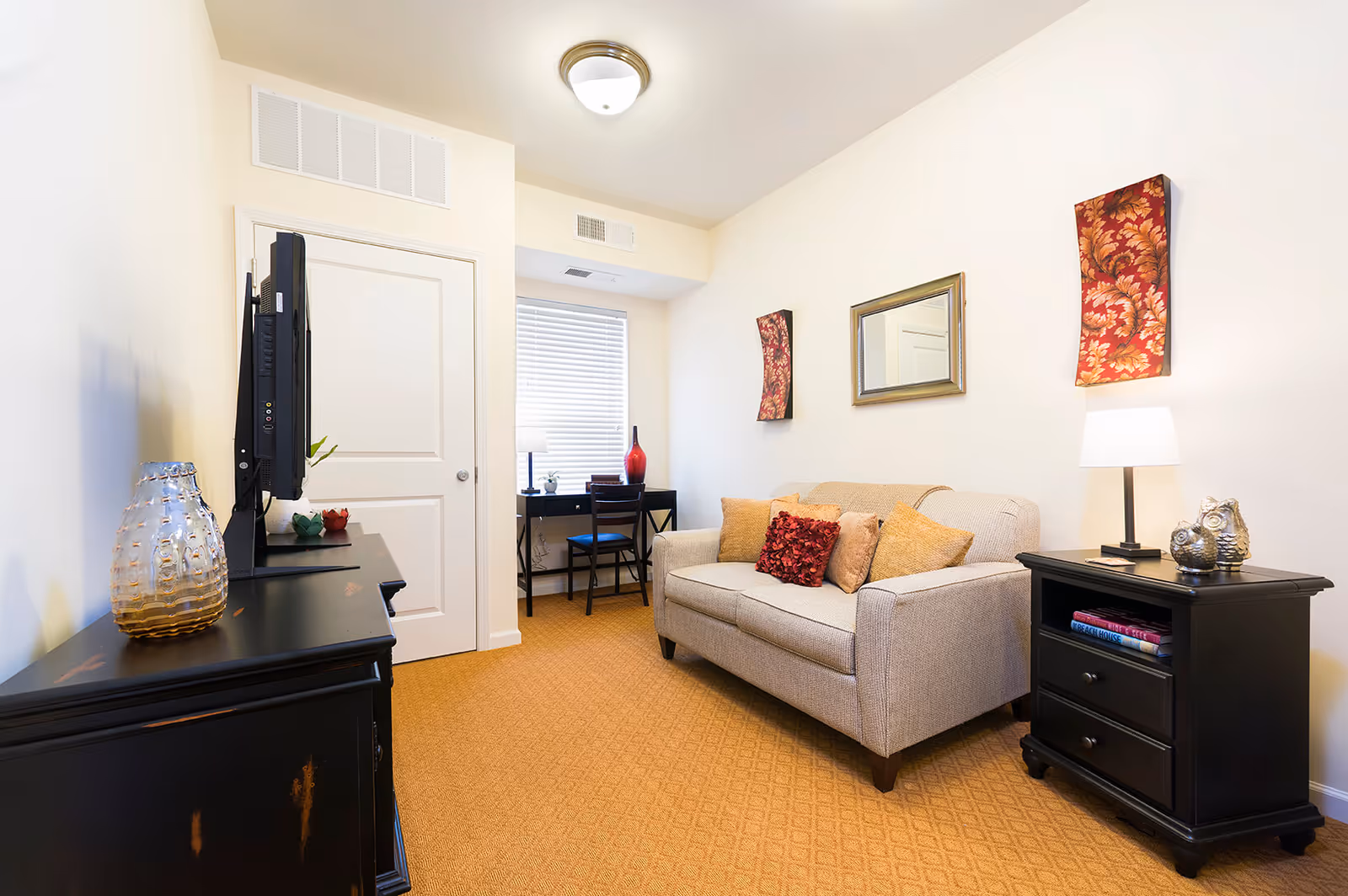 Small furnished living room with a beige sofa, decorative pillows, side tables, TV on a dresser, and a small desk by a window.