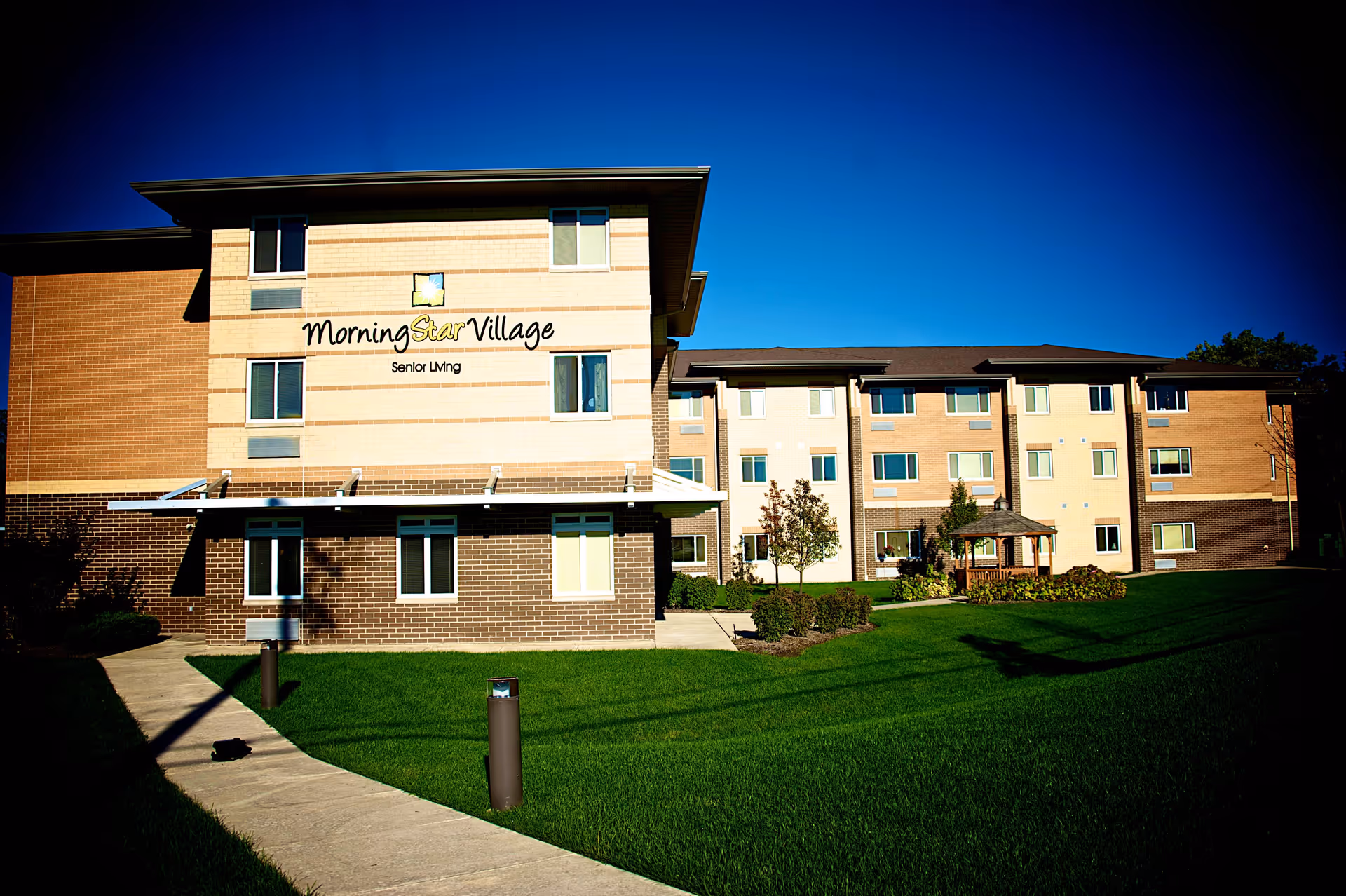 Exterior view of Morning Star Village senior living facility showing a multi-story building with beige and brown brick walls, several windows, a small gazebo, and a well-maintained green lawn under a clear blue sky.