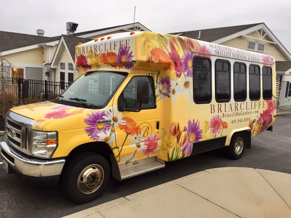 A yellow floral Briarcliffe Gardens shuttle bus parked in front of a senior living building.