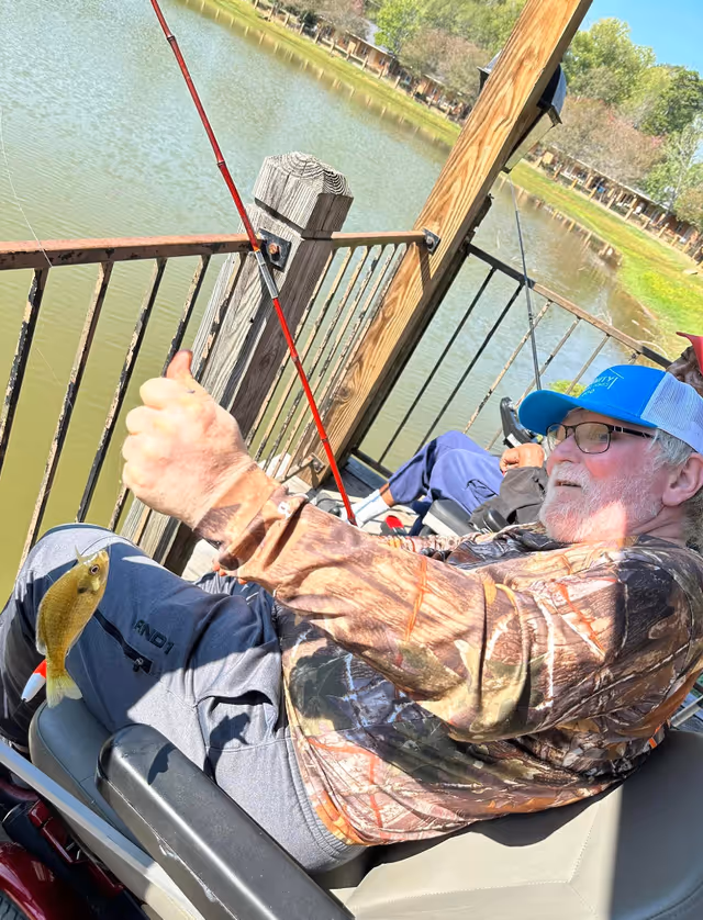 An elderly man wearing a camouflage jacket and blue cap is sitting on a bench by a wooden railing, holding a fishing rod with a small fish caught on the line. Another person is sitting beside him, also fishing, near a calm pond surrounded by trees and grass.