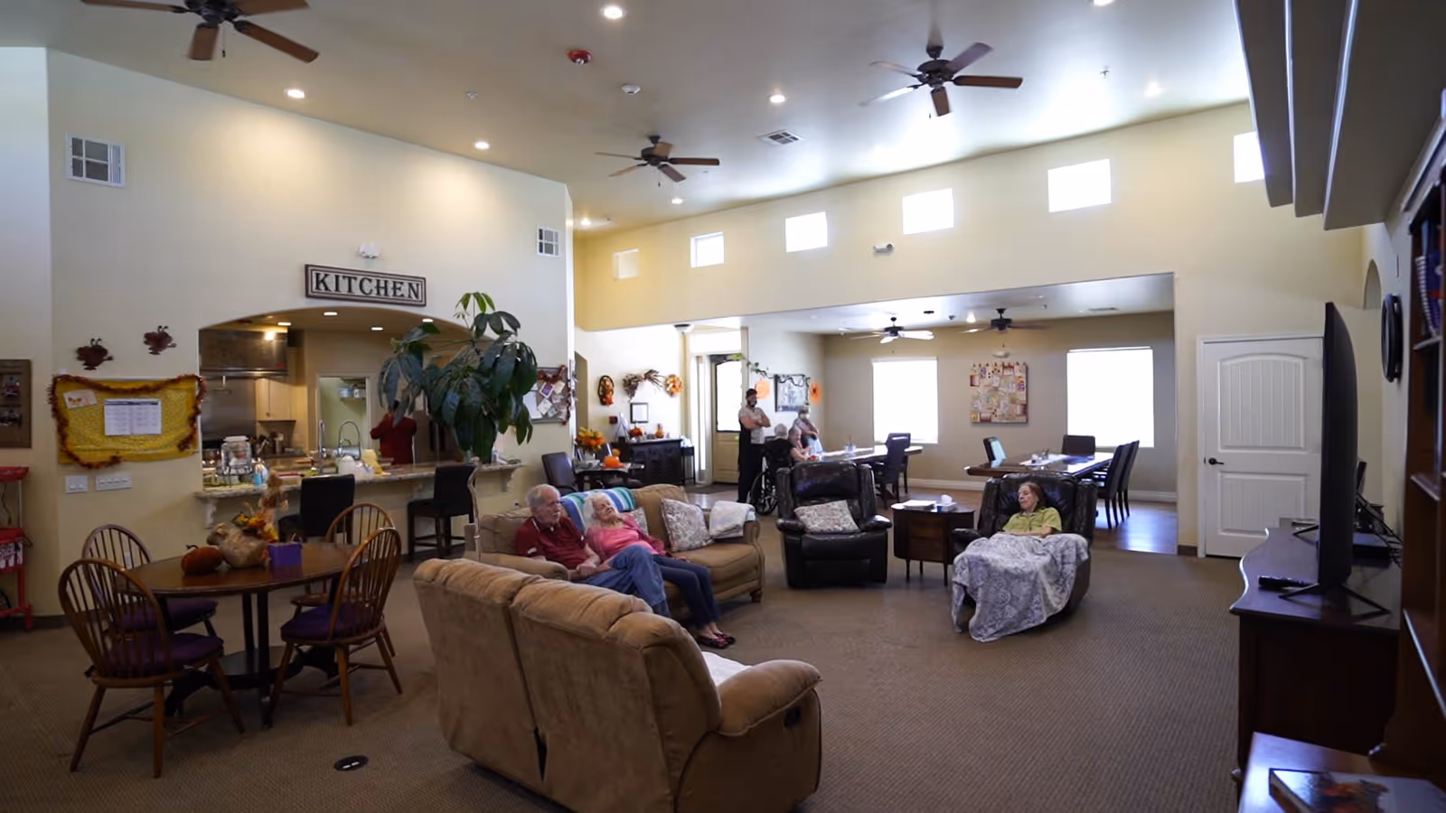 Open common room in a senior living facility with sofas and armchairs, dining tables, a kitchenette window labeled "KITCHEN", and residents seated around.