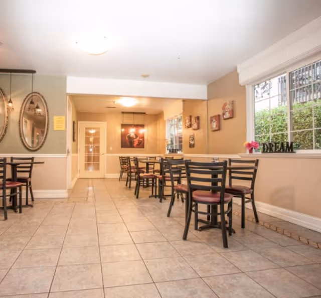 Bright communal dining room with tiled floor, multiple tables and chairs, wall art, and a window with decorative 'DREAM' letters.