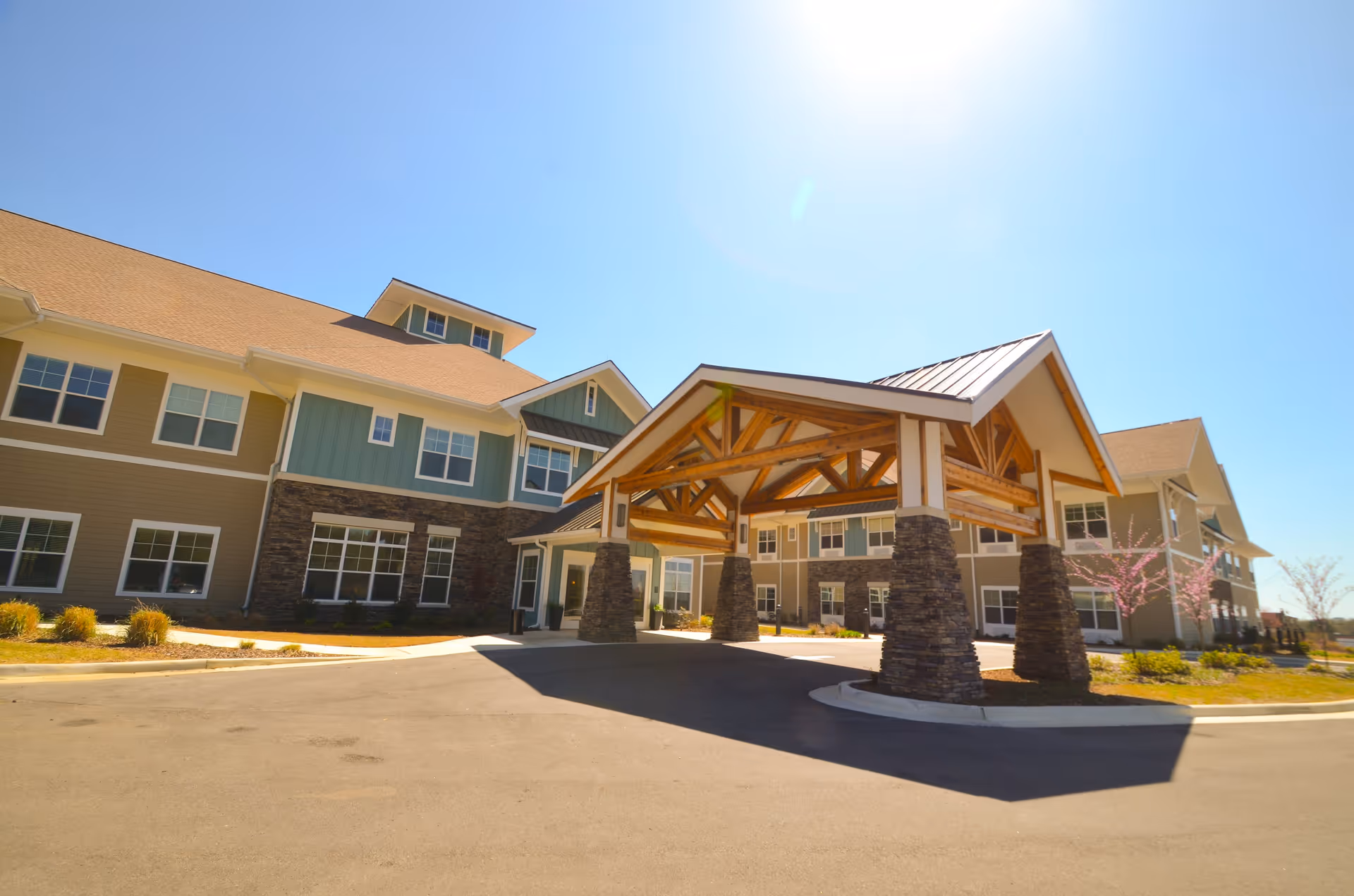 Exterior view of a senior living facility building with a large covered entrance supported by stone and wooden pillars under a clear blue sky.