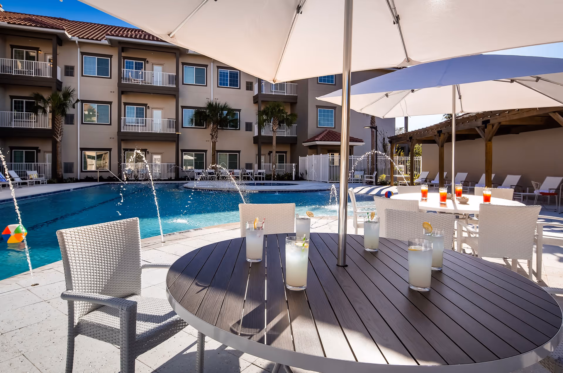 Outdoor pool area at Windsor Pointe of Jacksonville with a round table and white chairs under large umbrellas. Several glasses of lemonade and iced tea are on the tables. Water fountains spray into the pool, and a multi-story building with balconies and palm trees is visible in the background.