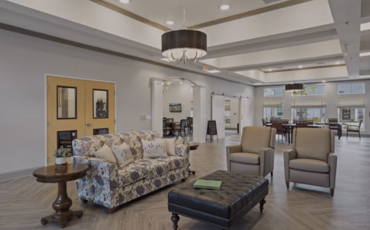Spacious communal living room with a patterned sofa, two armchairs, a tufted ottoman and side tables, with dining tables visible in the background under coffered ceilings.