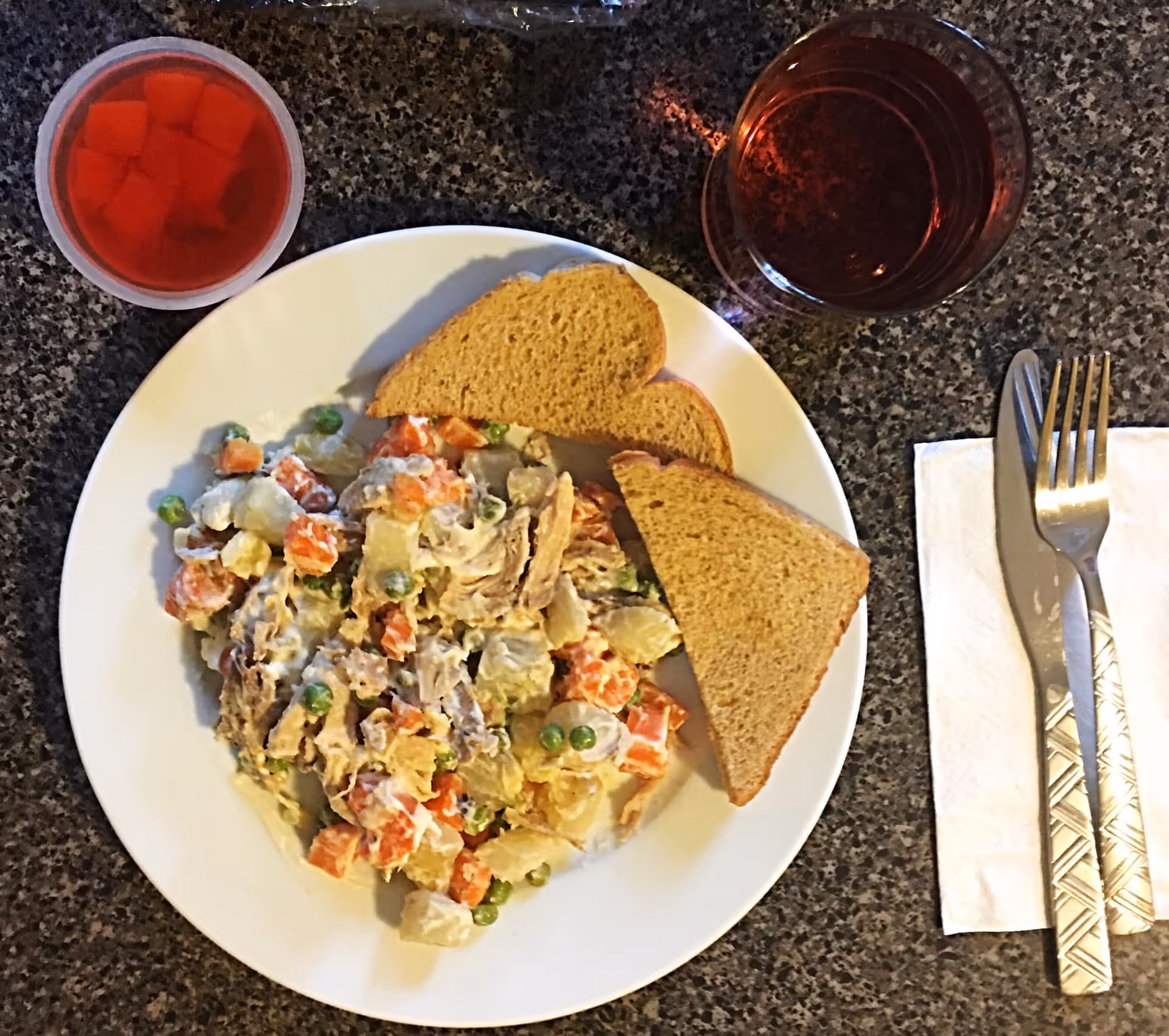 Top-down view of a plate with mixed potato-and-vegetable salad and two slices of bread, a cup of red gelatin, a beverage, and utensils on a napkin.