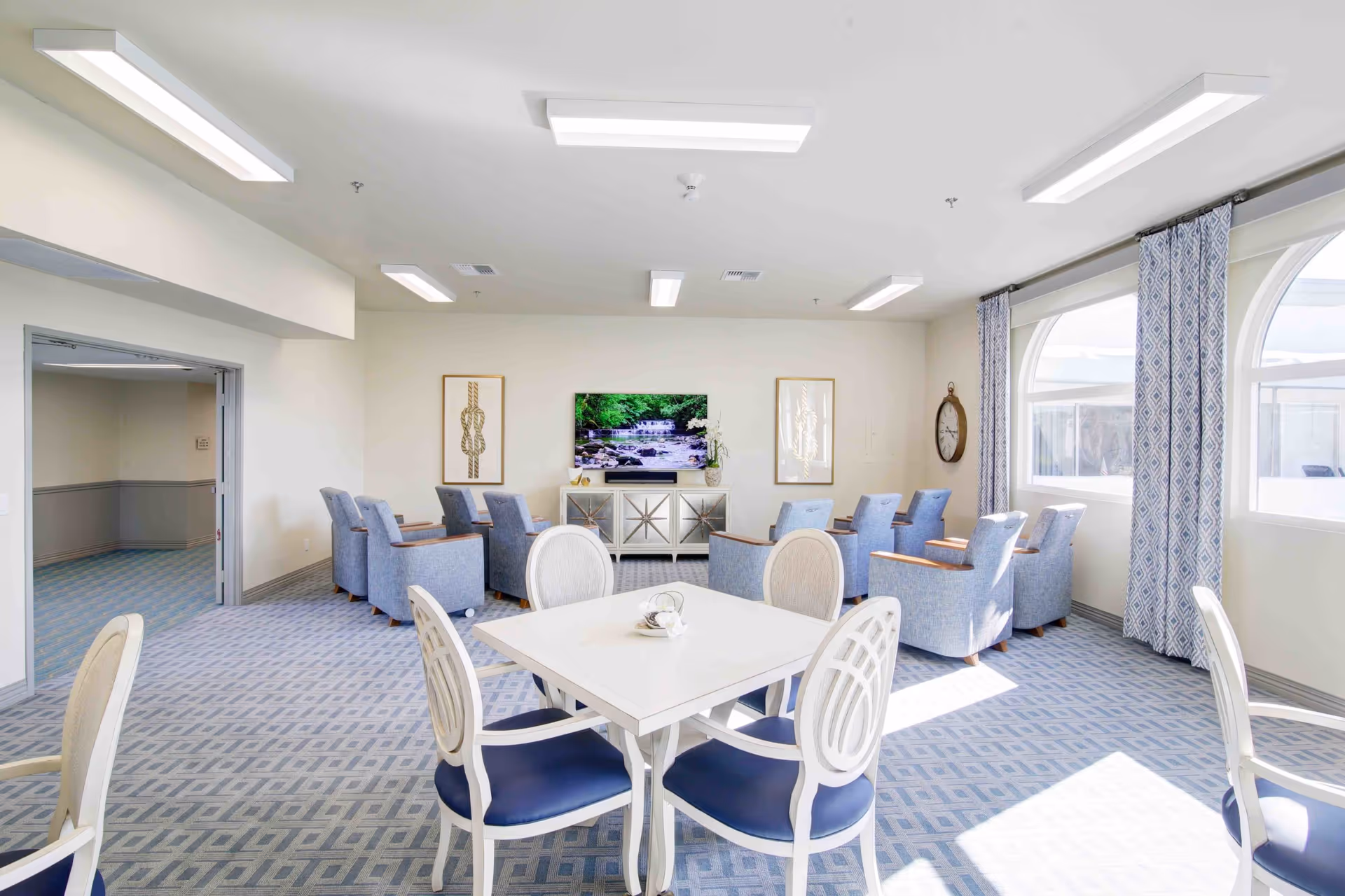 A bright and spacious senior living common area with a square white table and four white chairs with blue cushions in the foreground. In the background, there are eight blue armchairs arranged in two rows facing a flat-screen TV mounted on a white wall. The room has large arched windows with patterned blue curtains allowing natural light to fill the space. The floor is carpeted with a blue geometric pattern, and there are framed artworks and a wall clock on the walls.