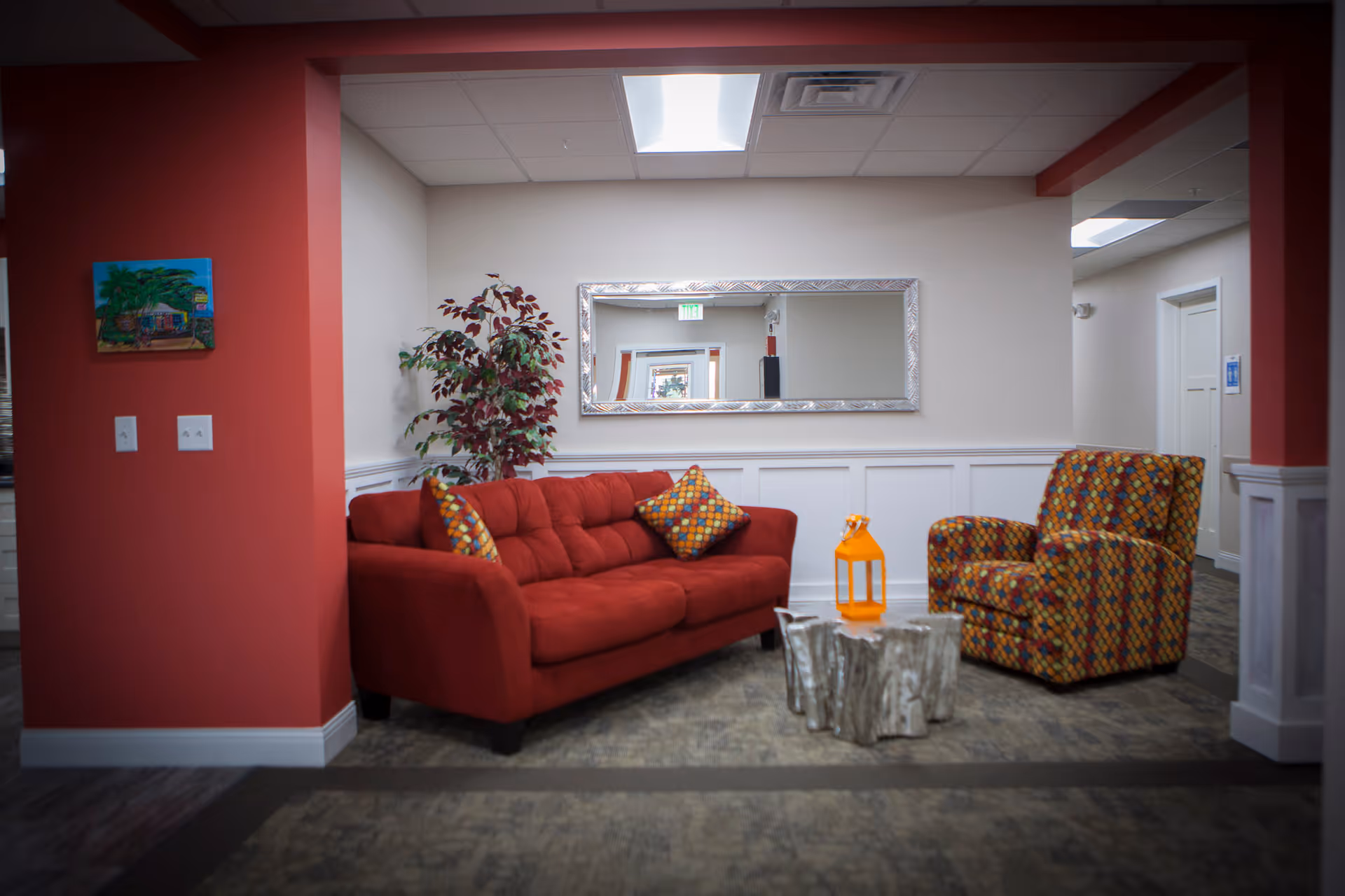 A cozy seating area with a red sofa, patterned armchair, decorative plant, and a large mirror on the wall.