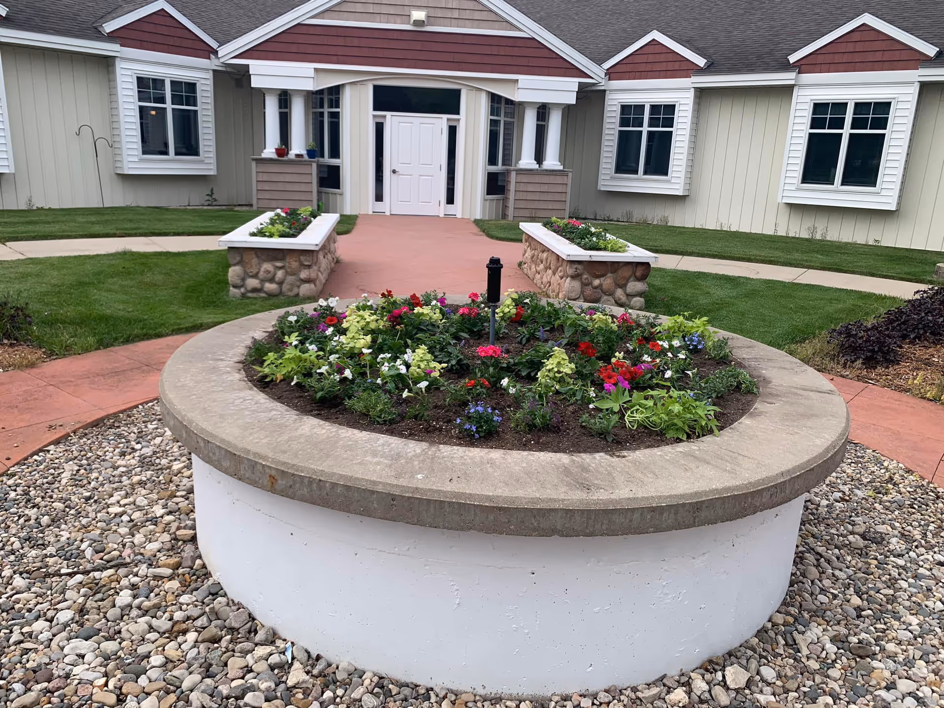 Circular raised planter filled with colorful flowers in a pebble courtyard leading to the entrance of a single-story building.