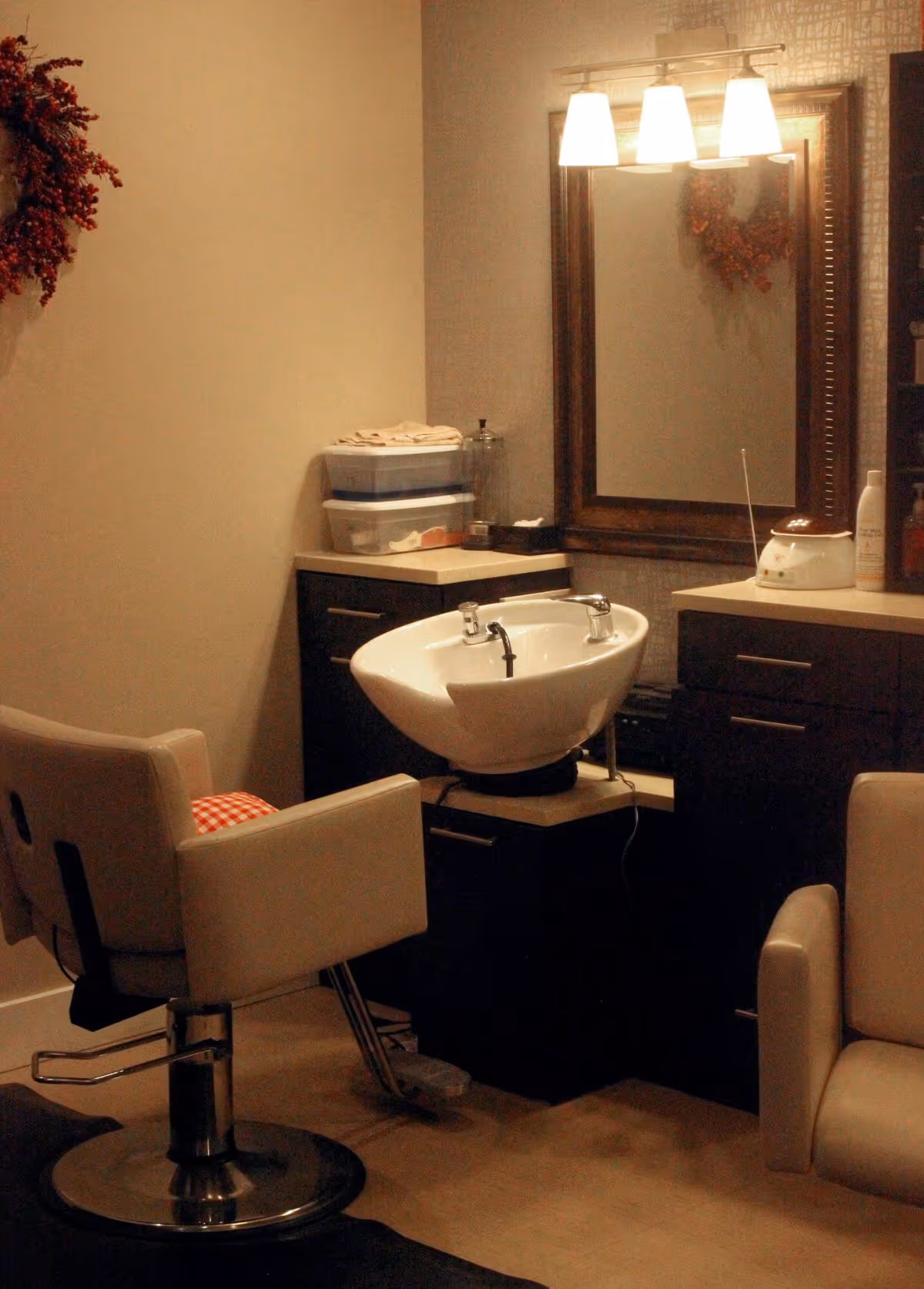 Interior view of a salon area with a white salon chair in front of a white hair washing sink. There is a large framed mirror on the wall with three lights above it. Dark cabinetry with countertop surrounds the sink, and various salon supplies are placed on the counters. A wreath decoration is partially visible on the wall.