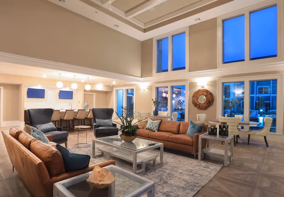 Spacious and well-lit living room area with high ceilings and large windows showing an evening sky. The room features two brown leather sofas, two dark armchairs, a glass coffee table with a plant centerpiece, side tables, and a rug. In the background, there is a kitchen area with a bar counter and four bar stools, illuminated by three pendant lights. A round decorative mirror hangs on the wall near a small dining table with four white upholstered chairs.