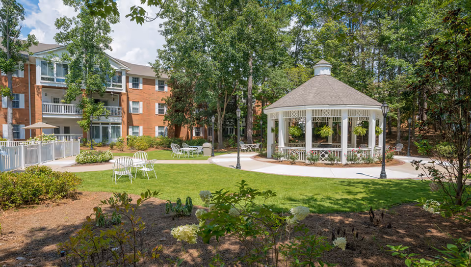 Sunlit courtyard featuring a white gazebo, lawn chairs, and a brick apartment building surrounded by trees.