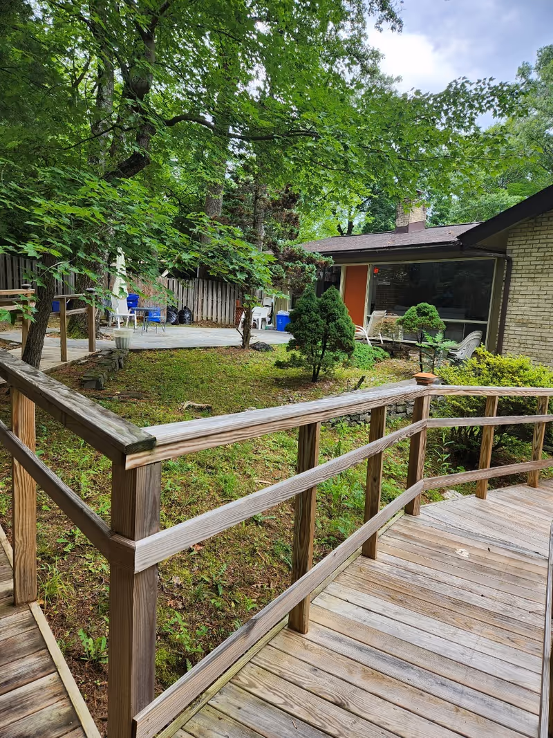 A wooden ramp with railings leading to a patio area surrounded by greenery and trees. The patio has chairs and tables, and a brick building with large windows is visible in the background.