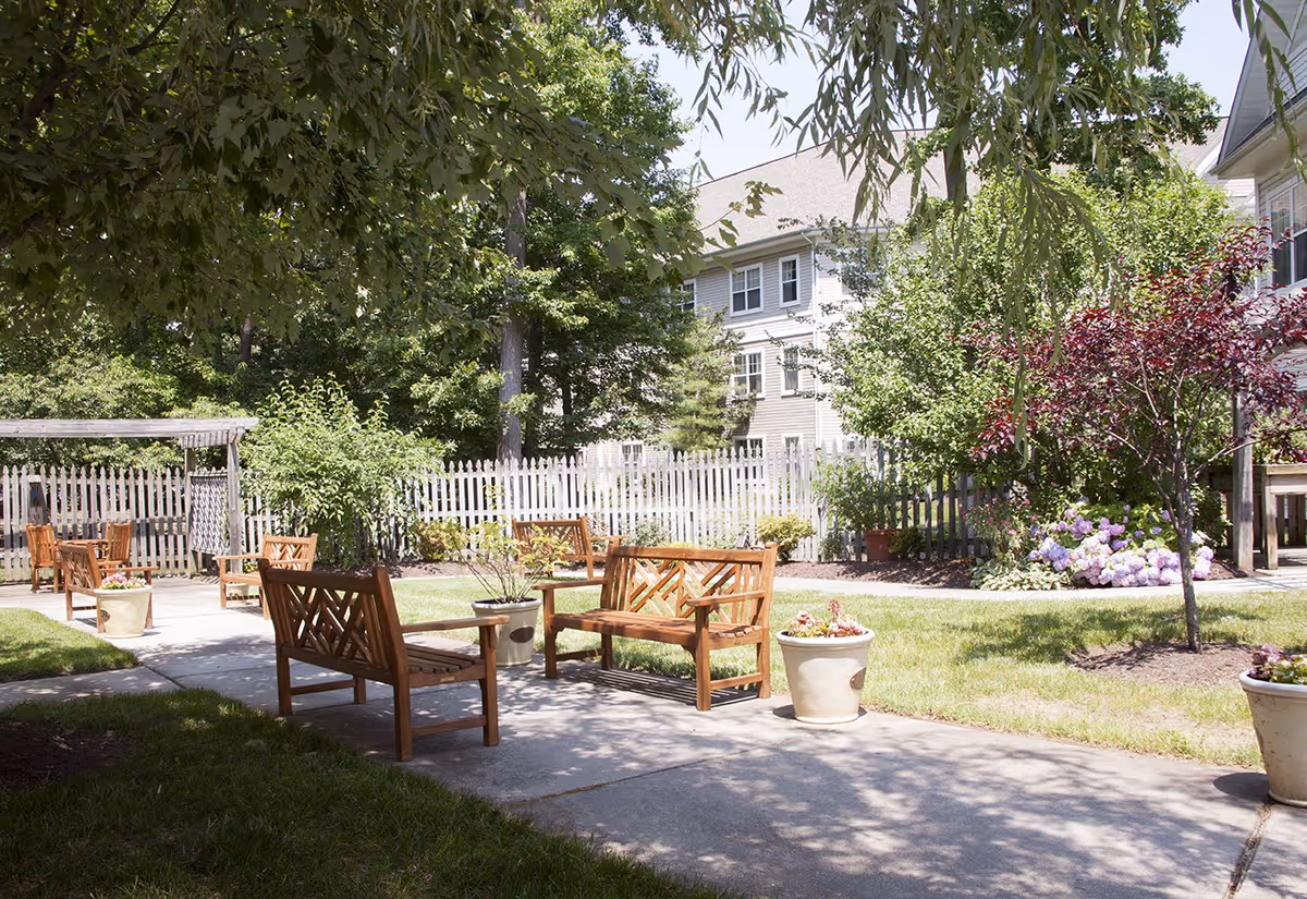 Outdoor garden area with wooden benches and potted plants along a concrete pathway, surrounded by green grass, trees, and a white picket fence. A multi-story building is visible in the background under a clear sky.
