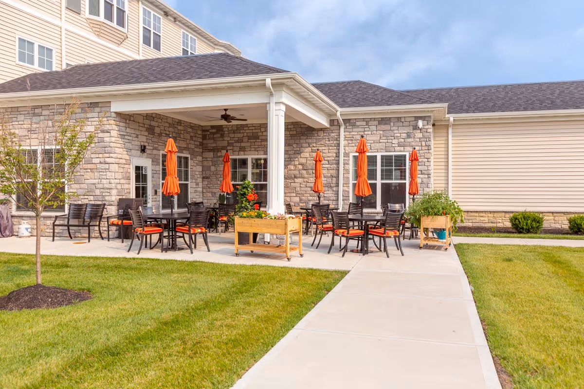 Outdoor patio area at StoryPoint Union with multiple tables and chairs featuring orange cushions and matching orange umbrellas. The patio is adjacent to a building with stone and siding exterior walls, and there is a concrete walkway leading to the patio surrounded by green grass and small plants.
