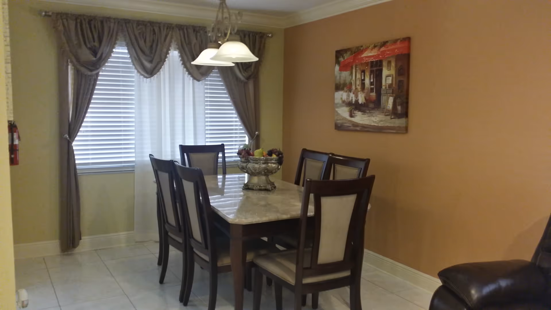Dining room with a rectangular marble-top table surrounded by six wooden chairs with cushioned backs and seats. A decorative bowl with fruit is placed in the center of the table. The room has beige walls, a window with blinds and curtains, a hanging light fixture above the table, and a framed painting on the wall.