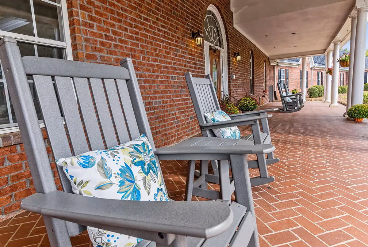Covered brick-front porch with gray rocking chairs and floral pillows, white columns, and potted plants along a tiled walkway.