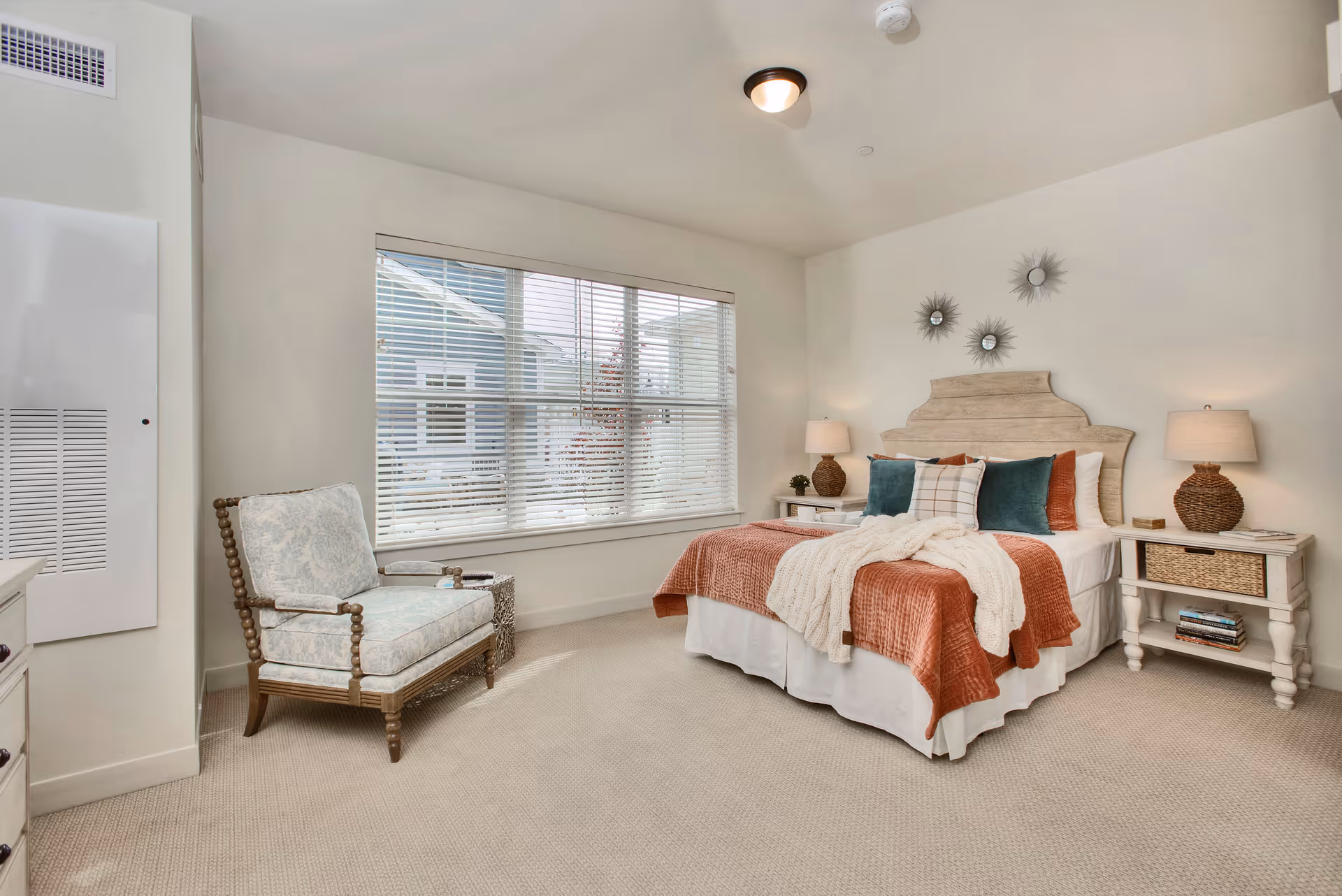 A bright bedroom with a large window covered by blinds, a bed with a wooden headboard, orange and white bedding, and multiple pillows. There are two bedside tables with lamps and decorative items, a cushioned armchair with wooden frame, and a small side table next to it. The walls are light-colored and decorated with three sunburst mirrors above the bed.