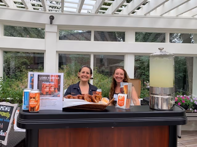 Two women standing behind a refreshment table with pretzels, canned drinks, and a large beverage dispenser filled with lemonade, inside a bright room with large windows and a glass ceiling.